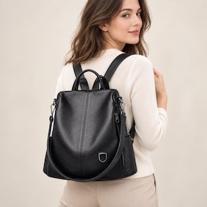 May include: A black backpack with a textured finish, featuring a top handle, adjustable shoulder straps, and a small silver-toned shield-shaped emblem on the front. The bag includes side pockets and a vertical seam detail down the centre front.