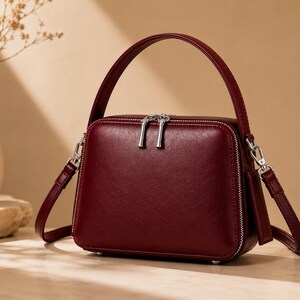 May include: A structured, deep red handbag with a smooth finish, featuring a top handle and a detachable shoulder strap. The bag has a rectangular shape with rounded corners, silver-toned metal zippers with dual pulls, and silver hardware attachments.