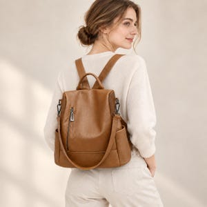 Puede incluir: Una mochila de color tostado de tamaño mediano hecha de material sintético liso. La bolsa cuenta con un bolsillo vertical con cremallera en la parte delantera, dos bolsillos laterales y correas ajustables con clips de metal oscuro.