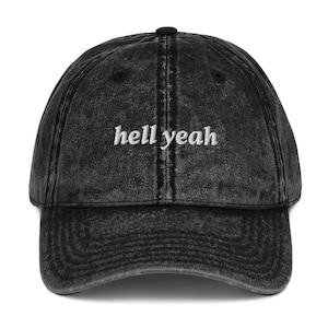 Pode incluir: Um boné de beisebol preto com efeito de lavagem ácida, apresentando o texto "hell yeah" bordado em fio branco nos painéis frontais. O boné possui uma copa estruturada, aba curva e um botão no topo central, com um acabamento de tecido desgastado.