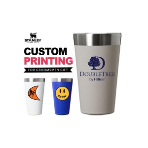 Puede incluir: Tres vasos Stanley con impresión personalizada. Un vaso grande de color beige presenta el logotipo de DoubleTree by Hilton. Un vaso blanco más pequeño muestra un gráfico de luna creciente naranja y un vaso azul muestra una cara sonriente amarilla con el texto drew.