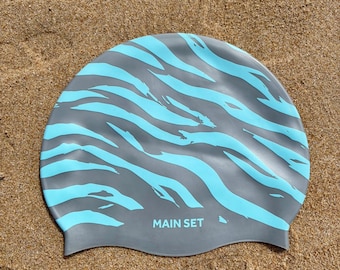 May include: A grey silicone swim cap featuring a light blue tiger stripe pattern across the surface. The text "MAIN SET" is printed in light blue capital letters near the bottom centre of the cap, which has a smooth, rounded, and flexible design.