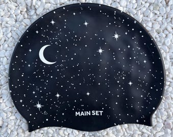 Peut inclure: Un bonnet de bain en silicone noir orné d'un motif céleste blanc représentant un croissant de lune et des étoiles de différentes tailles. Le texte MAIN SET est imprimé en lettres capitales blanches près du bord inférieur du bonnet.
