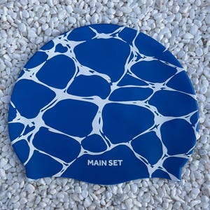 May include: A blue swim cap featuring a white, webbed, water-like pattern across the surface. The text "MAIN SET" is printed in white capital letters near the bottom centre of the cap. The design mimics the appearance of light reflecting on water.