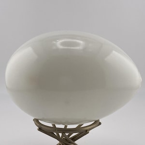 May include: A smooth, solid white egg-shaped object with a glossy finish, resting securely on a metallic, twisted wire stand that features a circular base and multiple intersecting prongs supporting the rounded form of the white egg.