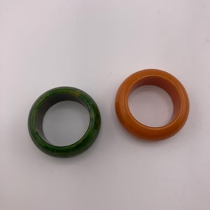 May include: Two circular rings are displayed. One ring features a mottled green and dark surface with a smooth finish. The second ring is a solid, opaque orange color with a rounded, polished exterior and a consistent circular shape.
