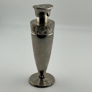 May include: A silver-toned metal perfume bottle with a tapered body and a circular base. The neck features an embossed floral pattern, and the top is finished with a flat, semi-circular cap. The surface has a polished, reflective metallic finish.