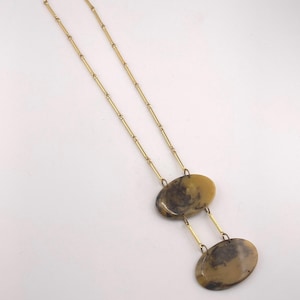 May include: A gold-toned necklace featuring a long chain with small cylindrical links. The pendant consists of two smooth, oval-shaped tan and dark brown marbled resin pieces connected by short gold-toned metal bars in a vertical arrangement.