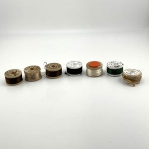 Puede incluir: Siete carretes de hilo de madera vintage dispuestos en una fila horizontal. Los carretes presentan varios colores, incluyendo marrón, beige, verde oscuro y blanco, con etiquetas de papel en los extremos que muestran texto impreso y valores numéricos.