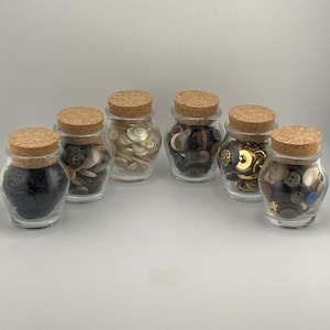 May include: Six clear glass jars with cork stoppers, each filled with a collection of assorted buttons. The jars contain various button colors including black, white, brown, and metallic gold, showcasing different sizes and shapes of sewing fasteners.