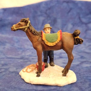 May include: A brown horse figurine with a black mane and tail, wearing a tan bridle and a green saddle blanket with yellow fringe. A small figure in a hat and dark clothing stands beside the horse on a white, snow-covered base with a red bucket.
