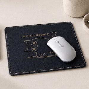 Peut inclure: Un tapis de souris rectangulaire noir avec des bords cousus blancs, orné d'un dessin au trait doré représentant un chat aux yeux écarquillés avec le texte IS THAT A MOUSE ?! et T.C. Une souris d'ordinateur blanche repose sur le tapis.