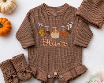 May include: A brown knitted long-sleeve romper with a ruffled hem and three buttons at the crotch. The chest features an embroidered garland of leaves, acorns, and a pumpkin with the name Olivia. Includes matching knit booties and a pom-pom beanie.