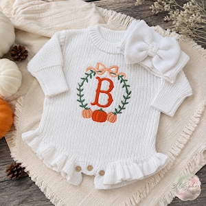 May include: A white long-sleeved knitted romper featuring a ruffled hem and a central embroidered design of a green wreath with orange pumpkins and a large orange letter B. A matching white knitted headband with a large bow is included.