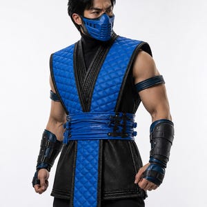 May include: A blue and black costume featuring a quilted blue vest with black trim, a blue face mask, matching blue arm guards with black straps, and a blue waist sash. The outfit includes a black high-neck base layer and black trousers.