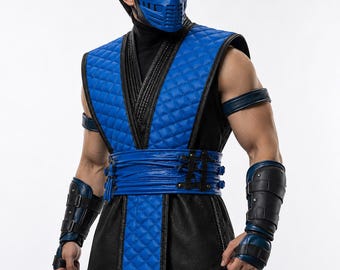 May include: A blue and black costume featuring a quilted blue vest with black trim, a blue face mask, matching blue arm guards with black straps, and a blue waist sash. The outfit includes a black high-neck base layer and black trousers.