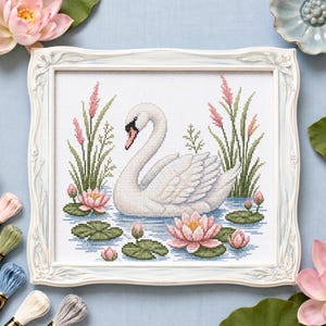 May include: A framed cross-stitch embroidery featuring a white swan swimming in blue water surrounded by green lily pads, pink water lilies, and tall pink-tipped reeds. The artwork is encased in a decorative, light-colored, ornate wooden frame.