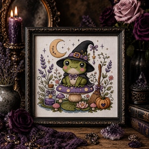 Puede incluir: Un bordado en punto de cruz enmarcado que muestra una rana verde con sombrero y capa de bruja negra, sentada sobre una seta morada. La escena incluye una luna creciente, una escoba, un frasco de poción, ramas de lavanda y una pequeña calabaza.