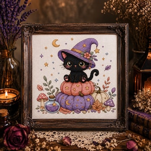 May include: A framed cross-stitch embroidery featuring a black kitten wearing a purple witch hat, sitting atop stacked pink and purple pumpkins. The design includes small stars, a crescent moon, mushrooms, and a potion bottle on a light fabric background.