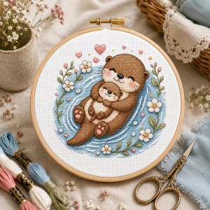 以下が含まれることがあります： 木製の円形刺繍枠に、青い水に浮かびながら小さな赤ちゃんカワウソを抱く茶色の母親カワウソのクロスステッチ模様が施されています。デザインは白い花、緑の葉、小さなピンクのハートで囲まれています。