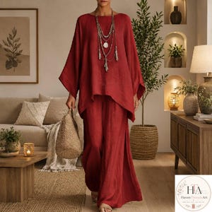 Puede incluir: Un conjunto de dos piezas de color rojo intenso que consta de una túnica holgada de manga larga y pantalones anchos. El conjunto se combina con un collar largo plateado de varias capas con dijes y un bolso de mano de paja tejida.