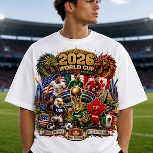 May include: A white t-shirt featuring a colorful 2026 World Cup graphic. The design includes the text 2026 WORLD CUP, USA, CANADA, MEXICO, THREE NATIONS, UNITED BY THE GAME, and ONE PASSION, alongside soccer players, flags, and mascots.