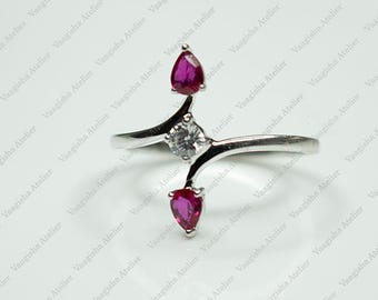 Puede incluir: Un anillo de tono plateado con diseño cruzado, que presenta una piedra central redonda y transparente flanqueada por dos piedras de color rosa intenso en forma de pera. El aro es liso y pulido, con una apariencia delicada y simétrica.