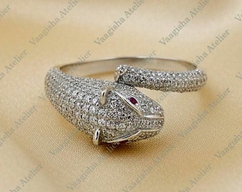 Puede incluir: Un anillo en forma de serpiente de tono plateado con una superficie pavé cubierta de numerosas piedras pequeñas y claras. La cabeza de la serpiente tiene una pequeña piedra roja como ojo y el cuerpo se enrolla para formar el anillo.