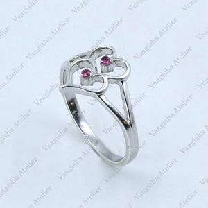 Könnte beinhalten: Ein silberfarbener Ring mit einem doppelten herzförmigen Design. Die Mitte jedes Herzens ist mit einem kleinen, runden, leuchtend rosa Edelstein akzentuiert. Die Ringschiene ist glatt und poliert und läuft in einer geteilten Form zusammen.