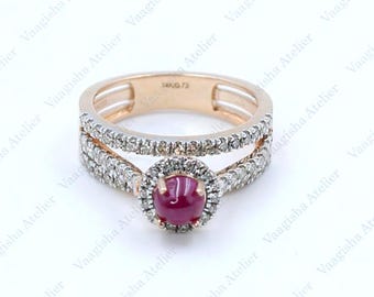 Puede incluir: Un anillo de oro rosa con un rubí rojo central en talla cabujón rodeado por un halo de pequeños diamantes blancos. La banda está dividida en dos filas, ambas incrustadas con diamantes blancos en pavé, creando un aspecto detallado.