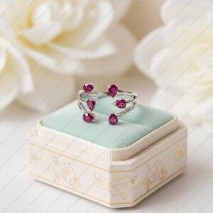 Peut inclure: Une bague ouverte en argent orn&eacute;e de quatre pierres pr&eacute;cieuses rouge ros&eacute; profond en forme de poire. L'anneau est divis&eacute; en deux sections distinctes, chacune portant deux pierres en forme de goutte aux extr&eacute;mit&eacute;s ouvertes, cr&eacute;ant un aspect sym&eacute;trique.