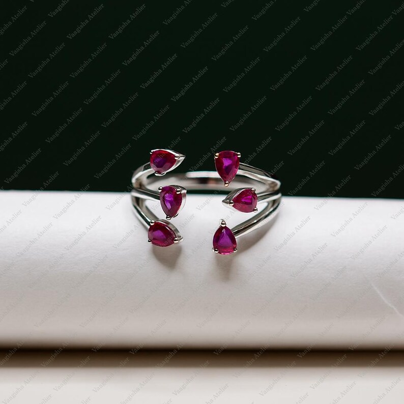 Peut inclure: Une bague ouverte argent&eacute;e orn&eacute;e de cinq pierres pr&eacute;cieuses rouge ros&eacute; en forme de poire. L'anneau se divise en plusieurs brins, chacun se terminant par une pierre taill&eacute;e en goutte, formant un agencement sym&eacute;trique avec une ouverture centrale.