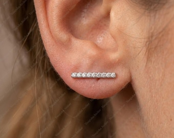 Peut inclure: Une boucle d'oreille de type grimpeur en ton argenté, présentant un design en barre horizontale orné d'une rangée de sept petites pierres claires taillées en rond. La boucle est placée le long du lobe de l'oreille, suivant sa courbe.