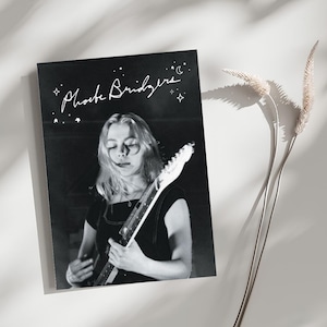 Puede incluir: Una impresión rectangular en blanco y negro con el retrato de una música tocando una guitarra eléctrica. El texto Phoebe Bridgers aparece en letra blanca en la parte superior, rodeado de pequeñas estrellas y una luna.
