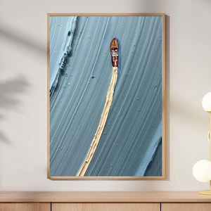 May include: A framed wall art print featuring an aerial view of a small brown wooden boat traveling across a textured light blue surface. The boat leaves a long, thick, cream-colored trail behind it, creating a contrast against the blue paint strokes.