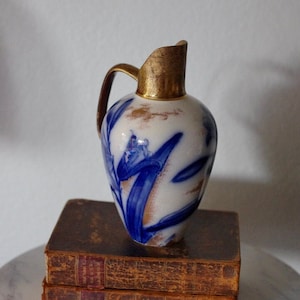 May include: A blue and white ceramic pitcher with gold accents and a floral design. The pitcher is sitting on top of a stack of old books.