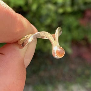 May include: A gold-toned ring featuring a smooth, curved band that splits into a V-shape at one end, terminating in a small, round, translucent orange gemstone set within a polished metal bezel.