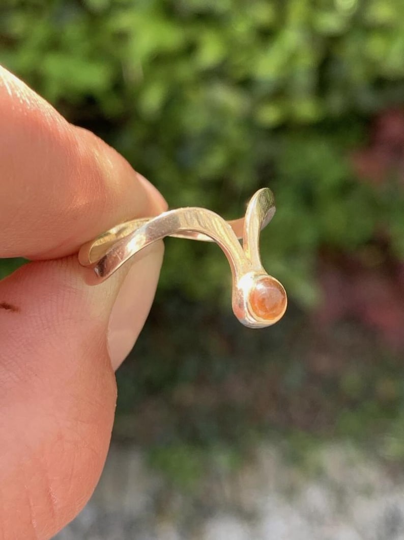 May include: A gold-toned ring featuring a smooth, curved band that splits into a V-shape at one end, terminating in a small, round, translucent orange gemstone set within a polished metal bezel.