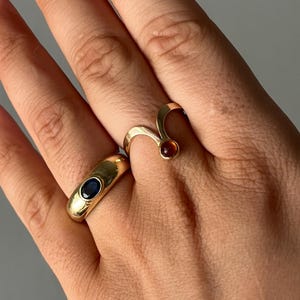 May include: Two gold rings are displayed on a hand. One ring is a thick band with a dark blue stone. The other ring has a curved design with a small, round, reddish-brown stone. The rings are set against a neutral background.