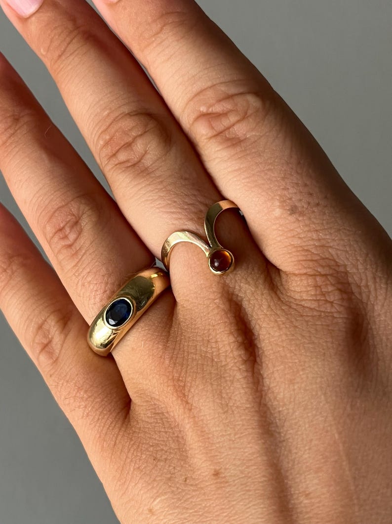 May include: Two gold rings are displayed on a hand. One ring is a thick band with a dark blue stone. The other ring has a curved design with a small, round, reddish-brown stone. The rings are set against a neutral background.