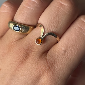 May include: Two gold rings are displayed on a hand. One ring is a thick band with an oval blue gemstone. The other ring is a curved design with a small, round, orange gemstone. The rings are set against a neutral background.