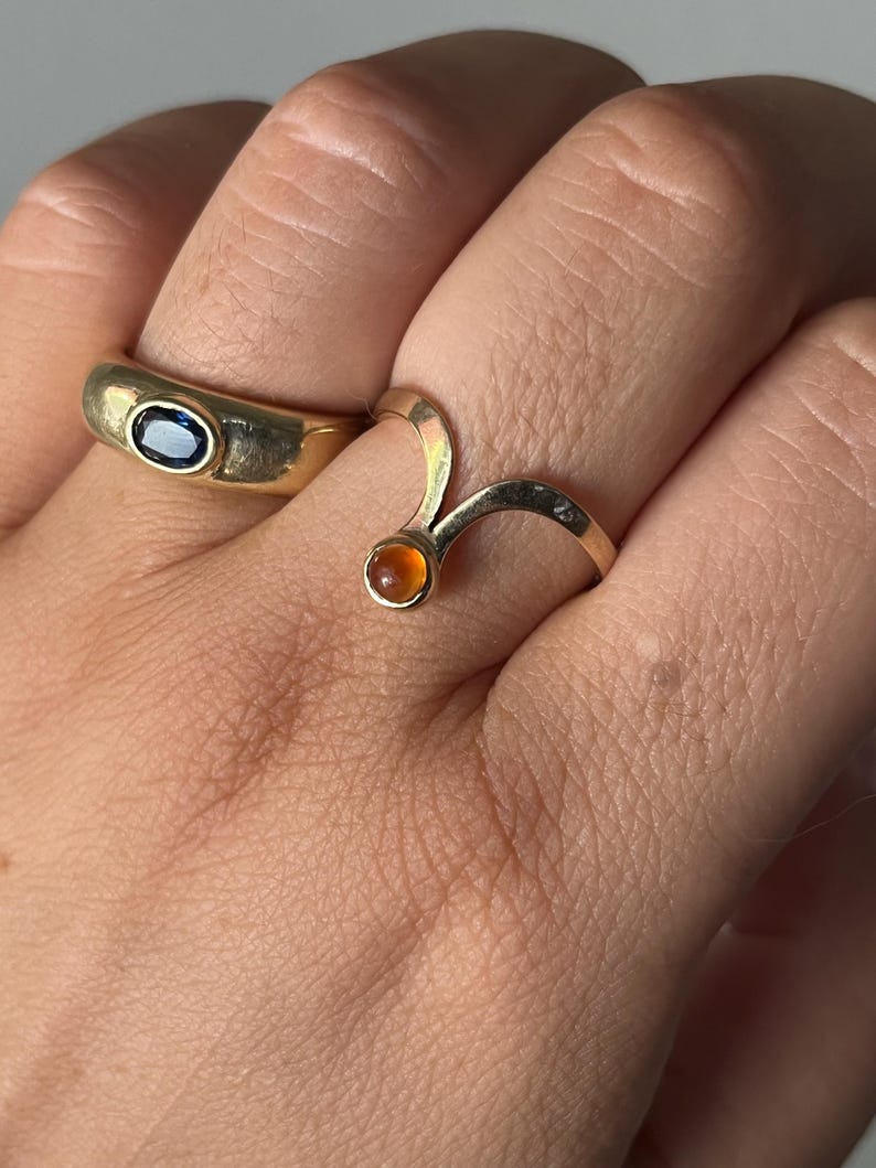May include: Two gold rings are displayed on a hand. One ring is a thick band with an oval blue gemstone. The other ring is a curved design with a small, round, orange gemstone. The rings are set against a neutral background.
