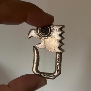 May include: A silver-toned metal pendant shaped like an eagle head with a dark circular inlay for the eye. The lower section is a U-shaped loop with stamped text that reads MEXICO and 925, indicating the metal composition and origin.