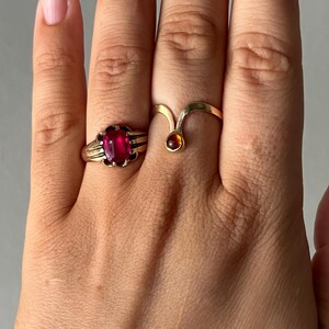 May include: Two gold rings are displayed on a hand. One ring features a large, oval, red gemstone set in a detailed gold band. The other ring has a unique, curved design with a small, round, amber-colored stone.
