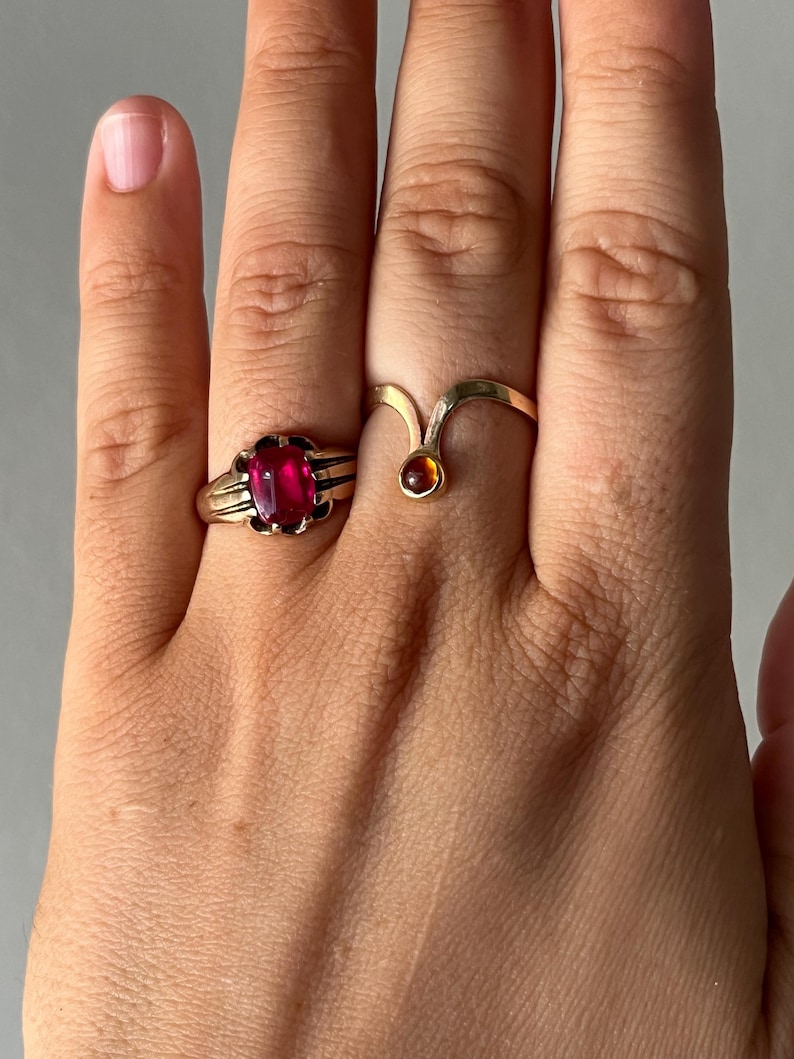 May include: Two gold rings are displayed on a hand. One ring features a large, oval, red gemstone set in a detailed gold band. The other ring has a unique, curved design with a small, round, amber-colored stone.