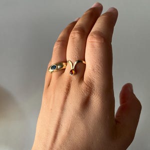 May include: A hand wearing two gold rings. One ring has a blue oval stone, and the other has a small, round, orange stone. The rings are set against a neutral background.