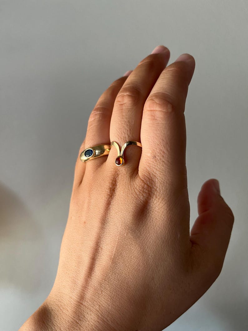 May include: A hand wearing two gold rings. One ring has a blue oval stone, and the other has a small, round, orange stone. The rings are set against a neutral background.