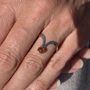 May include: A silver ring with a gold setting and a small, round, amber-colored gemstone. The ring has a unique, curved design. The ring is on a person's finger.