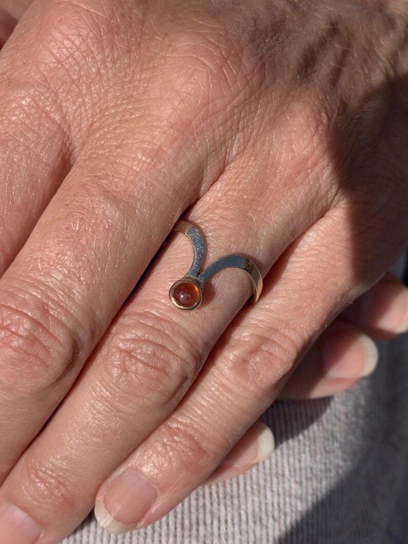 May include: A silver ring with a gold setting and a small, round, amber-colored gemstone. The ring has a unique, curved design. The ring is on a person's finger.