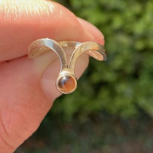 May include: A gold-toned ring featuring a V-shaped band that splits into two arms, meeting at a central point where a small, round, amber-colored gemstone is set. The metal has a smooth, polished finish, and the design creates a distinct arching shape.
